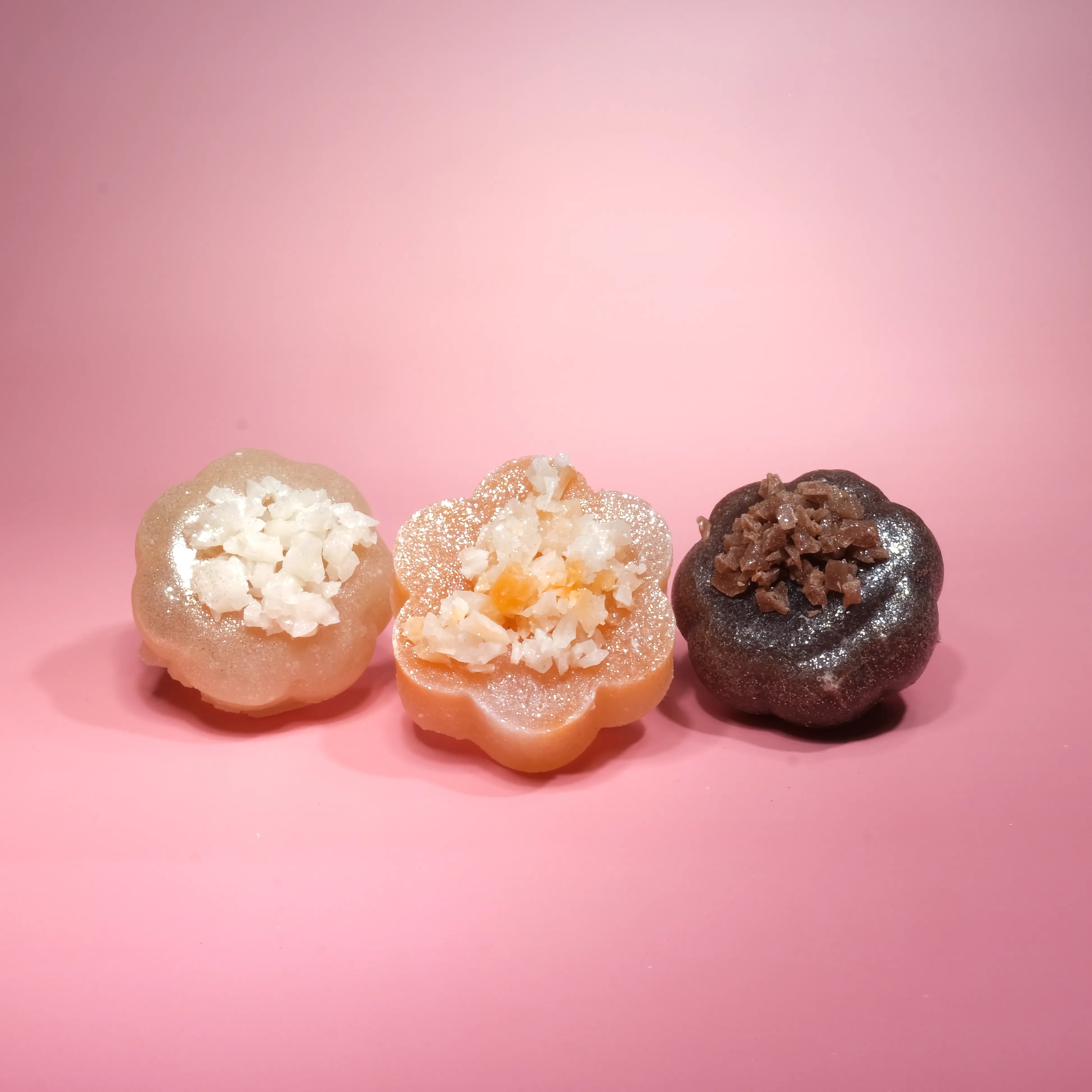 Three handcrafted crystal candies displayed side by side on a soft pink background, featuring creamy beige, warm peach, and dark cocoa-toned pieces topped with sparkling sugar crystals.