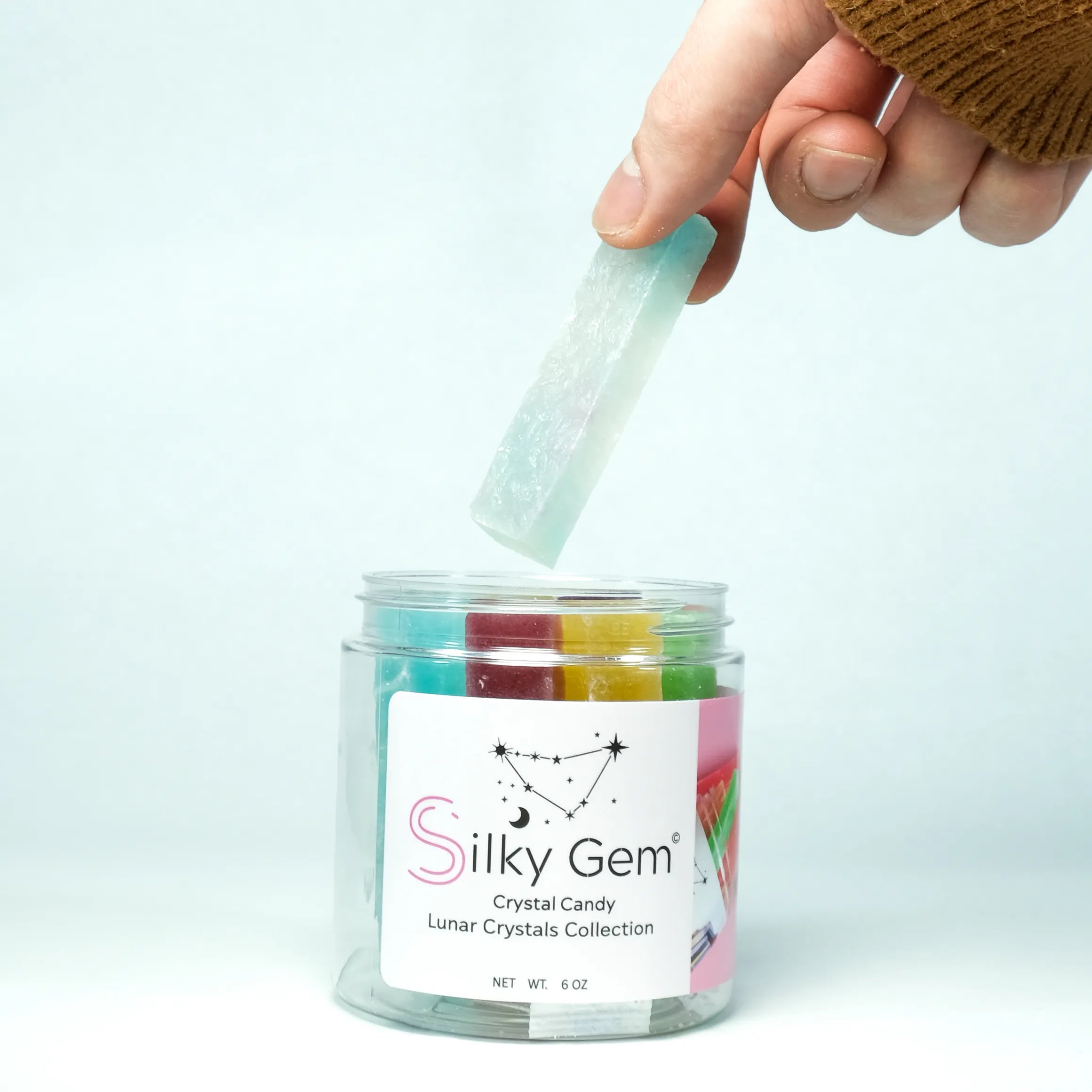 Hand lifting a translucent crystal candy from a Silky Gem Lunar Crystals jar, revealing colorful crystal candies inside, symbolizing Lunar New Year celebration.