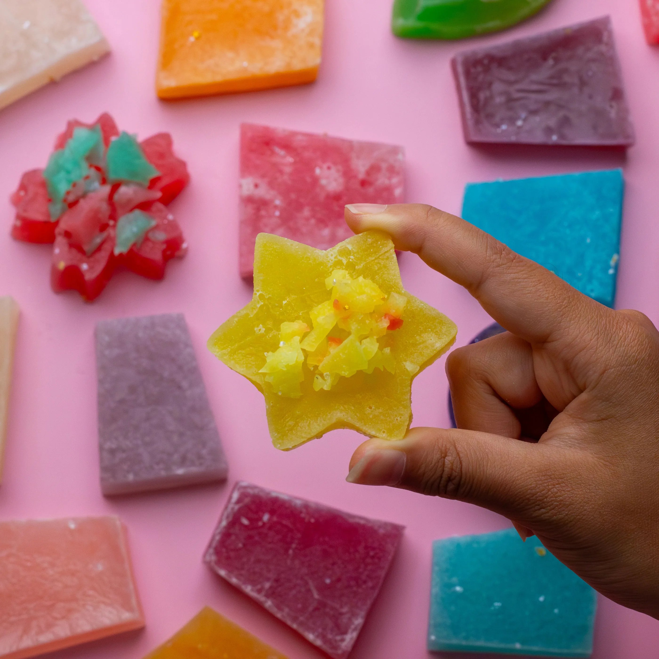 Colorful gummy candy shaped like a star held by a hand against a pink background with more gummy candies.