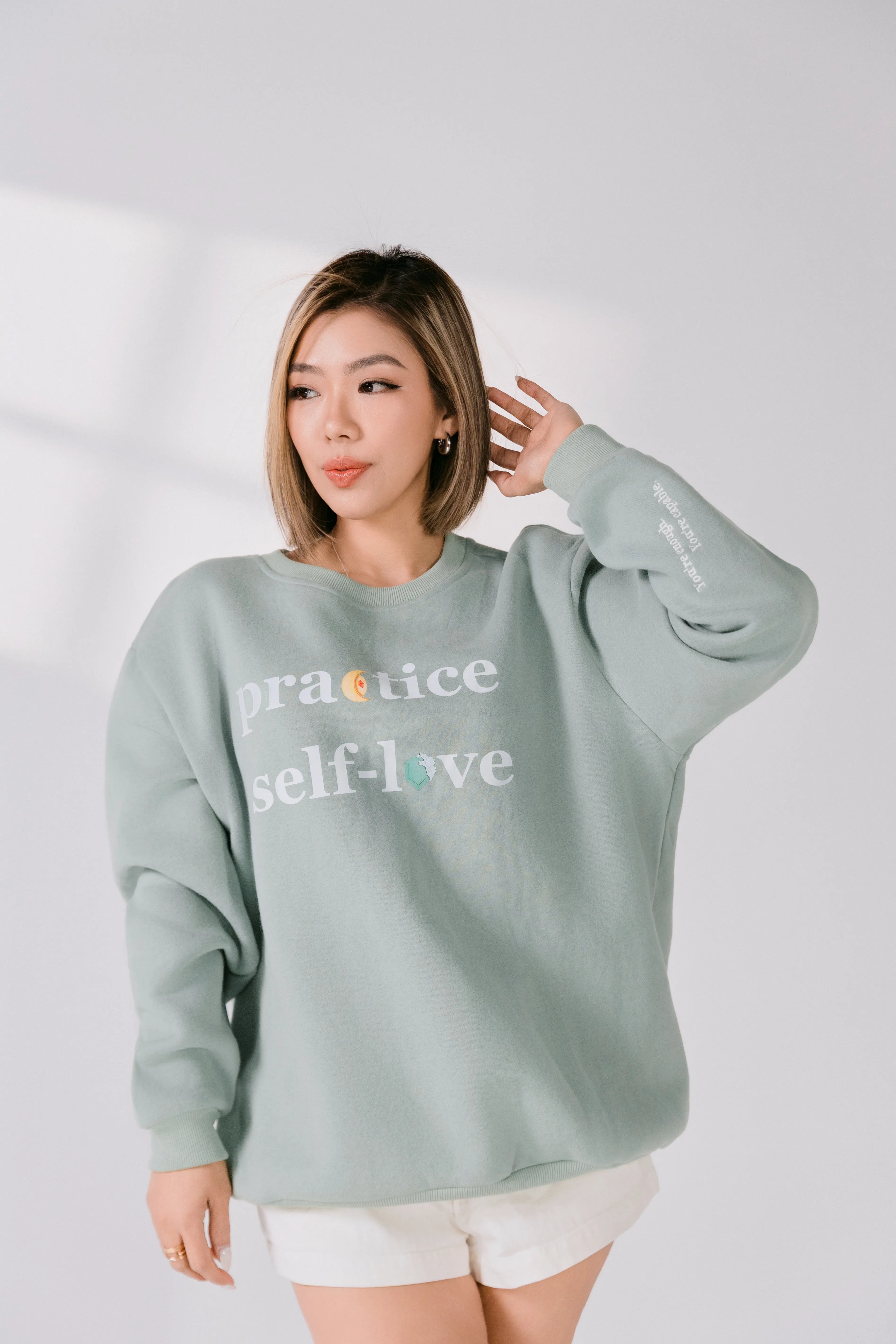 Model adjusting hair while wearing sage green motivational sweatshirt.