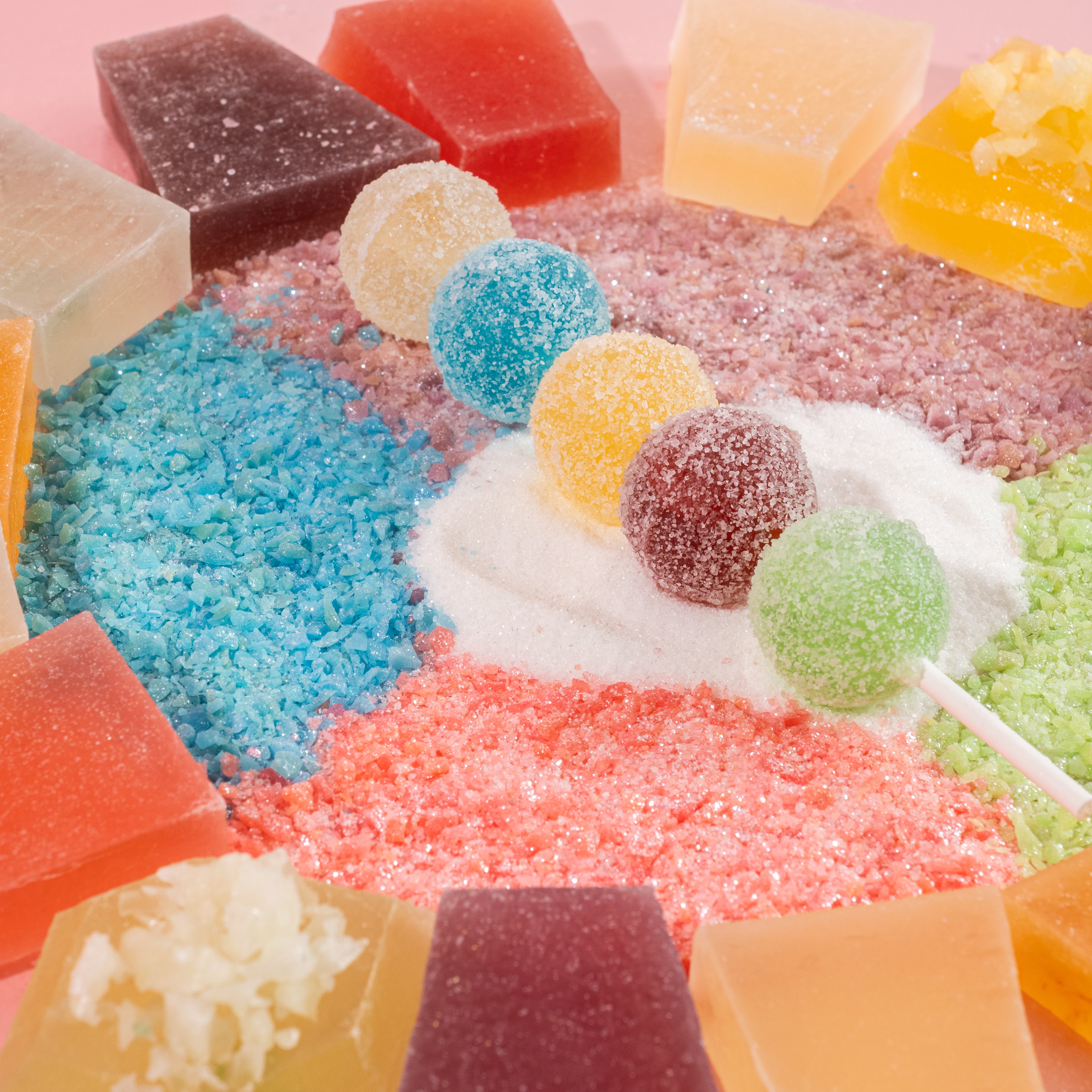 Artistic arrangement of vibrant Silky Gem crystal candies, including sphere and cube-shaped pieces in various colors, displayed on a bed of granulated sugar on a pink background.