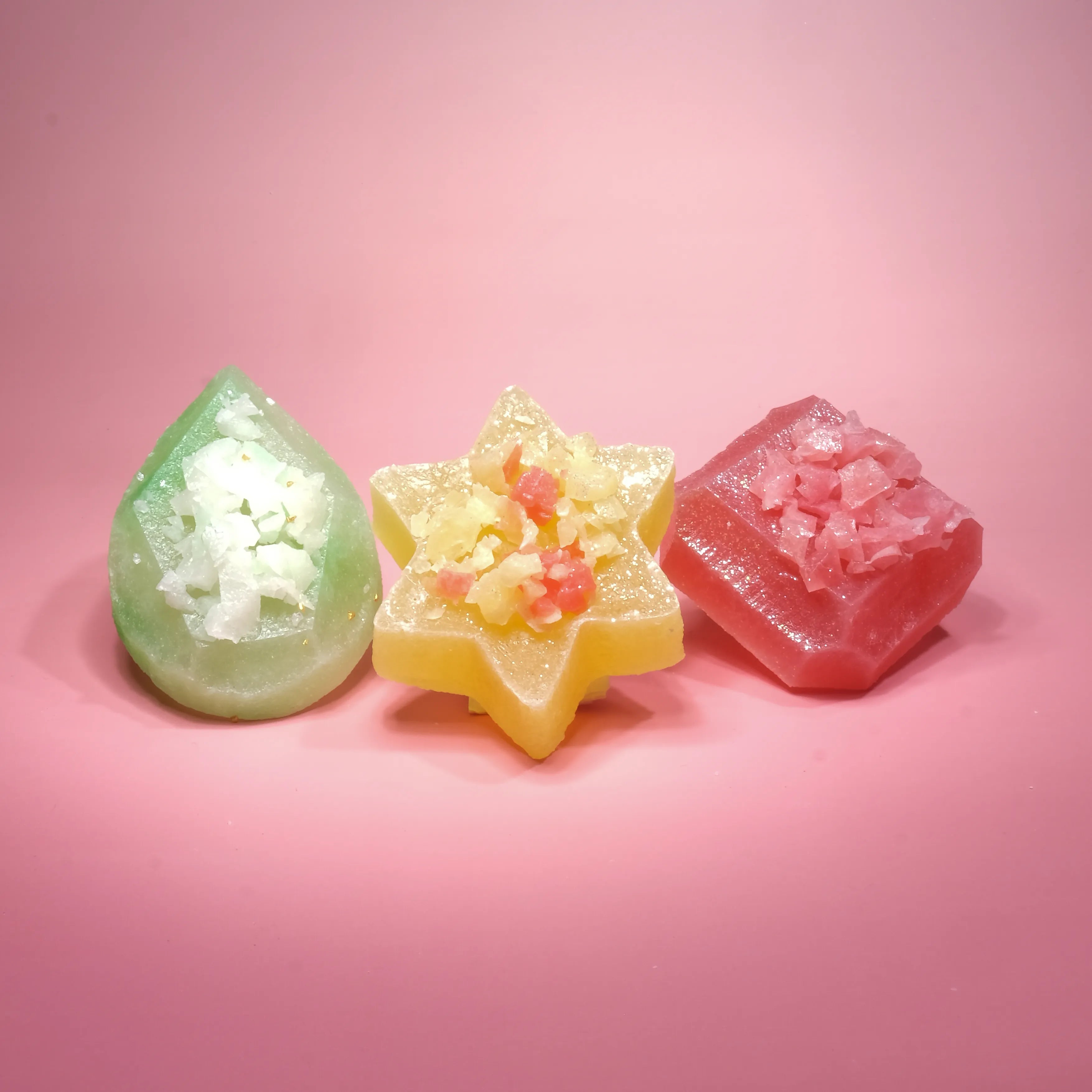 Three crystal candies on a pink background featuring a green teardrop candy, a yellow star-shaped candy, and a pink gemstone-shaped candy, each topped with shimmering sugar crystals.