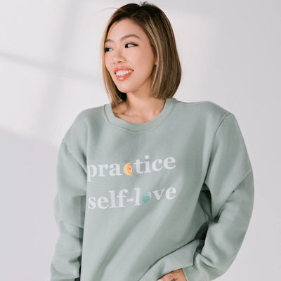 Person wearing a light green sweatshirt with 'practice self-love' text on a plain background