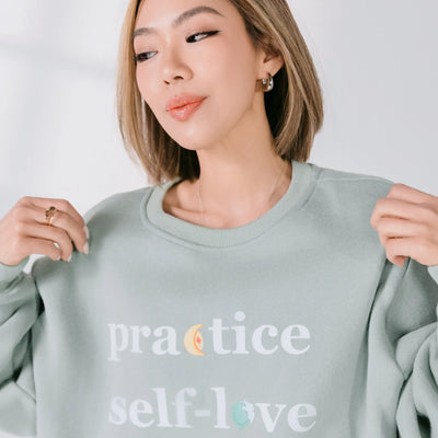Person wearing a light gray sweatshirt with 'practice self-love' text on a plain background