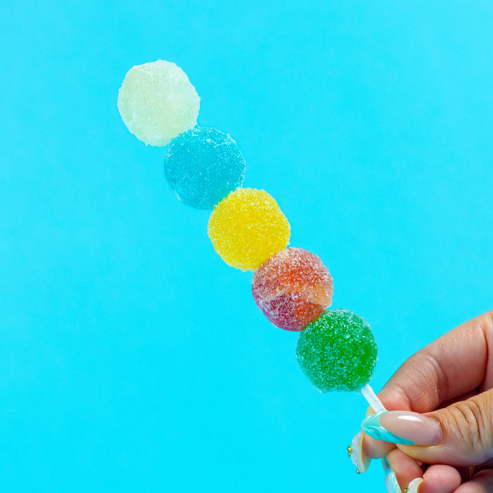 Hand holding a Sizzle Pop skewer featuring five colorful, crystal-cut gummy candies against a bright blue background.